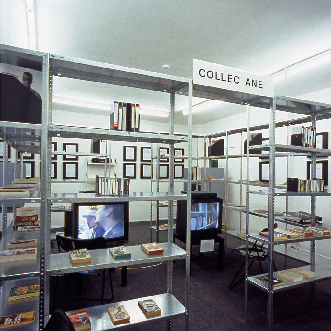 蕾妮·格林，《进口/出口放克办公室》，1992-93，混合媒介，金属层架、书籍、杂志、报纸、收藏品、BETA录像带、25小时数字化影像、两台显示器、一个影像投影、磁带、声音、亚克力标志、四个木制结构、四个架子上的亚克力标志、四台磁带播放器、三十个印有橡皮章文字的木牌。展览现场，科隆Galerie Christian Nagel，1992. 摄影：Andrea Stappert.