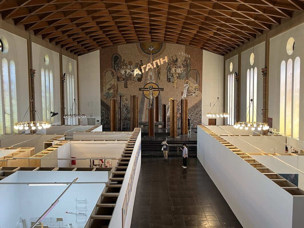埃森圣格特鲁德教堂展览现场. 可见一半区域用作埃森美院工作室，主祭坛镶嵌画上投影为舍伊拉·卡梅里奇作品《无条件的爱》（Agape，2026），前方竖立的教堂长椅为纳桑·图尔作品《升起》（Elevation, 2026）. 